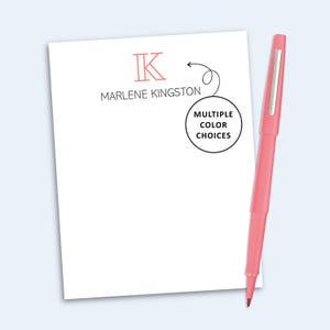 May include: A white notepad featuring a pink letter K, the name MARLENE KINGSTON in black text, and a circular graphic with an arrow pointing to the text MULTIPLE COLOR CHOICES. A pink pen with a silver clip is positioned to the right of the notepad.