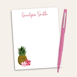 May include: A white rectangular notepad featuring the name Gracelynn Smithe in pink script at the top and a watercolor illustration of a pineapple with pink hibiscus flowers at the bottom left, accompanied by a pink ballpoint pen.
