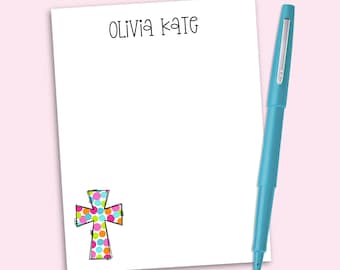May include: A white rectangular notepad featuring the name OLIVIA KATE in black lettering at the top and a colorful polka dot cross graphic in the bottom left corner, accompanied by a bright blue pen positioned vertically along the right side.