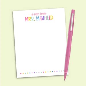 May include: A white rectangular notepad featuring the text a note from MRS. MAYFIELD in colorful lettering at the top and a row of small multicolored dots at the bottom, accompanied by a solid pink retractable ballpoint pen positioned vertically.