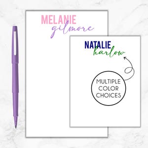 May include: Two white notepads with personalized names. The top notepad has the name "MELANIE gilmore" in pink and purple lettering. The bottom notepad has the name "NATALIE harlow" in blue and green lettering. A circle with the text "MULTIPLE COLOR CHOICES" is below the name.