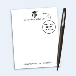 May include: A white notepad featuring a black caduceus symbol, the text Dr. Charlene Miller, DPT, and contact information at the bottom. A black pen rests beside the notepad, which includes a circular graphic with the text MULTIPLE COLOR CHOICES.