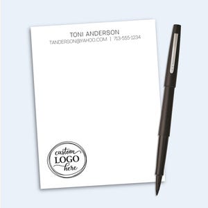 May include: A white rectangular notepad featuring the printed name TONI ANDERSON, an email address, and a phone number at the top, with a circular custom logo here graphic at the bottom left, accompanied by a black pen with a silver clip.