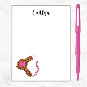 May include: A white notepad with the name "Caitlyn" printed at the top. The notepad features a cartoon illustration of a leopard print hairdryer with pink accents and a cord.