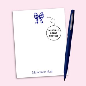 May include: A white notepad featuring a blue and white gingham bow illustration at the top, the name Makenzie Hall printed in blue at the bottom, and a circular graphic with the text MULTIPLE COLOR CHOICES, accompanied by a dark blue pen.