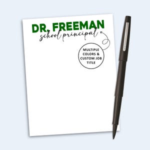 May include: A white notepad with the green text DR. FREEMAN and the black script text school principal. A circular graphic contains the text MULTIPLE COLORS & CUSTOM JOB TITLE with an arrow pointing to the text. A black pen is placed beside it.