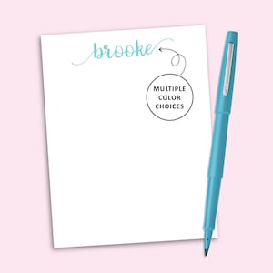 May include: A white notepad with the name brooke printed in light blue script at the top. A black circular graphic with an arrow pointing to the name contains the text MULTIPLE COLOR CHOICES. A light blue felt tip pen lies beside the notepad.