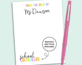 May include: A white notepad with the text FROM THE DESK OF Ms. Dawson in colorful letters and school counselor in black and colorful script. A black circle contains the text MULTIPLE JOB TITLES AVAILABLE with an arrow pointing to the title. A pink pen.