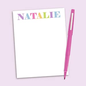 May include: A white rectangular notepad featuring the name NATALIE printed in multicolored pastel letters at the top, accompanied by a bright pink felt-tip pen positioned vertically along the right side of the paper sheet.