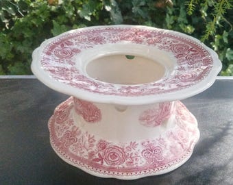 May include: A ceramic fondue pot base featuring a cream-colored background with a detailed red transferware pattern of floral motifs and landscape scenes. The circular stand has a wide top rim, a hollow center, and scalloped edges.