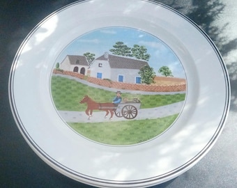 May include: A white ceramic dinner plate featuring a central illustration of a person driving a horse-drawn cart on a path in front of a stone house. The plate has a thin black circular rim detail and the name Laplau printed at the bottom.