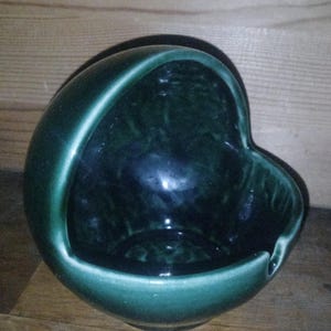 Könnte beinhalten: Ein dunkelgrüner Keramik-Aschenbecher mit einem einzigartigen herzförmigen Design. Der Aschenbecher hat eine glänzende Oberfläche und einen gebogenen Griff. Der Innenraum ist ein dunklerer Grünton, und der Rand ist heller grün.