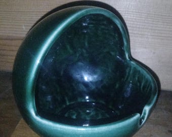 May include: A dark green ceramic ashtray with a unique heart-shaped design. The ashtray has a glossy finish and a curved handle. The interior is a darker shade of green, and the rim is a lighter green.