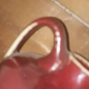 Può includere: Un'anfora in ceramica rossa con un bordo bianco e un manico. L'anfora ha una finitura liscia e lucida.