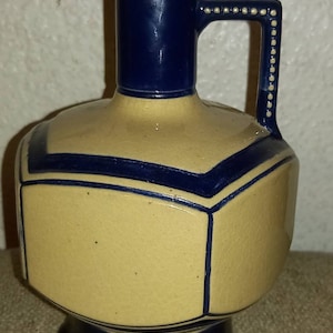 Könnte beinhalten: Vintage Keramikkrug mit cremefarbenem Körper und marineblauen Akzenten. Der Krug hat ein geometrisches Design, einen marineblauen Griff und einen quadratischen Sockel. Der Griff ist mit kleinen weißen Punkten verziert.