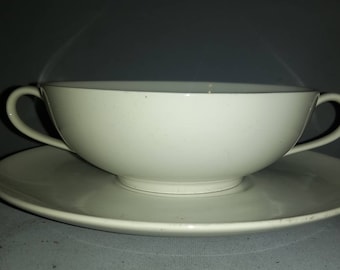 May include: A white ceramic soup bowl with two handles and a matching saucer. The bowl is filled with a steaming liquid.