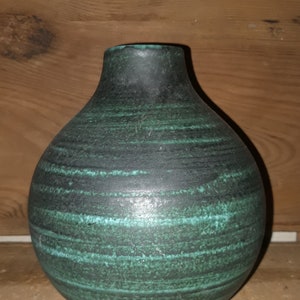 Puede incluir: Un jarrón de cerámica verde oscuro con cuerpo redondo y cuello estrecho. El jarrón tiene un patrón texturizado a rayas.