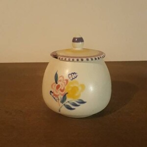 Könnte beinhalten: Eine weiße Zuckerdose aus Keramik mit Deckel. Die Schale ist mit einem Blumendesign in Rosa, Gelb und Blau verziert. Der Deckel hat einen kleinen Griff und einen dekorativen Rand.