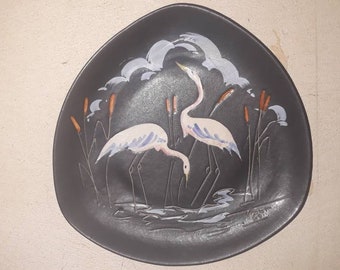 May include: A black ceramic plate with a white and blue design of two birds standing in a marsh. The birds are stylized and have long necks and legs. The marsh is depicted with reeds and water.