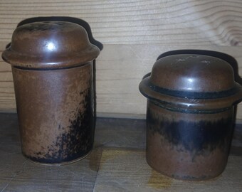 May include: Two brown ceramic salt and pepper shakers with lids. The shakers have a cylindrical shape with a dark brown and black mottled glaze. The lids are a darker brown and have a rounded shape.