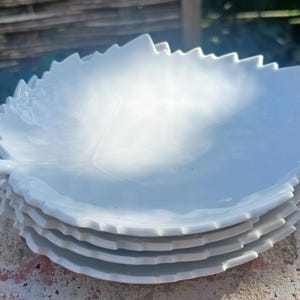 Puede incluir: Una pila de seis platos blancos de cerámica con forma de hoja. Cada plato tiene un patrón detallado de venas de hoja y un borde dentado. Los platos están apilados sobre una superficie de ladrillo, con un fondo borroso de madera y vegetación.