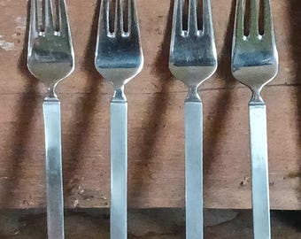 May include: Four silver forks with a rectangular handle design. The forks are arranged in a row against a wooden background. The tines of the forks are visible, and the handles are a lighter color than the tines.