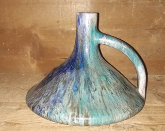 May include: A blue, green, and white ceramic vase with a unique, curved handle. The vase has a wide, flared base and a narrow neck.