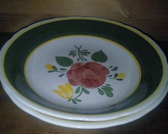 May include: A stack of ceramic plates with a floral design. The plates have a white center with a red rose and green leaves. The rim is dark green with a yellow border. The plates are stacked on top of each other.