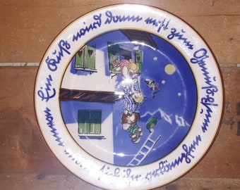 May include: A blue and white ceramic plate with a whimsical illustration of two children climbing a ladder to a house. The plate has a yellow rim with the text "Ein biß'l Sonnenschein, macht's Leben schön" in German.