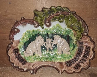 May include: A ceramic dish shaped like a leaf with three elephants in a forest scene. The dish is decorated with green, brown, and white glaze. The text "Alsens Oeffnungs Uhr 18 Uhr" is written on the dish.