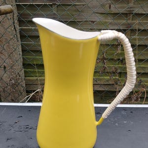 Könnte beinhalten: Eine leuchtend gelbe Kanne mit weißem Innenraum und einem geflochtenen weißen Griff. Die Kanne hat eine geschwungene Form und einen kleinen Ausguss. Der Griff besteht aus einem dicken, gewebten Material. Die Kanne ist wahrscheinlich aus Keramik oder Emaille.