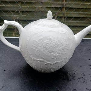 Può includere: Una teiera in porcellana bianca con corpo rotondo e coperchio. La teiera presenta disegni floreali e grappoli d'uva in rilievo. Ha un manico curvo e un beccuccio. Il coperchio è sormontato da un piccolo pomello decorativo.