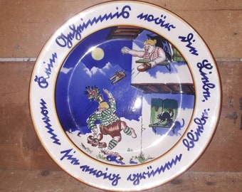 May include: A blue and white ceramic plate with a whimsical scene of two people on a balcony. The plate has a yellow rim and blue lettering around the edge that reads "Einz'n Oig'ninn, im moin'g g's'inn, sin'n Limbl'n, blimm'n."