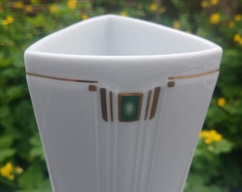 May include: A white triangular vase with a gold trim and a green square accent. The vase has vertical lines and a geometric design. The background is blurred with green foliage and yellow flowers.