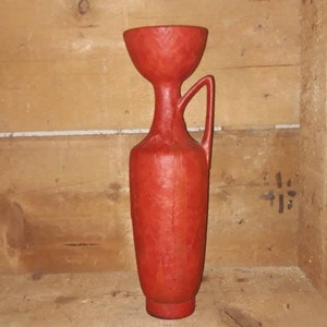 Può includere: Un vaso in ceramica rosso con una superficie testurizzata e un manico. Il vaso ha un'ampia apertura e una base stretta.