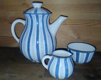 May include: A white ceramic tea set with blue vertical stripes. The set includes a teapot with a lid, a creamer, and a small bowl. The stripes vary in shade, creating a visually appealing design. The set is placed on a wooden surface.