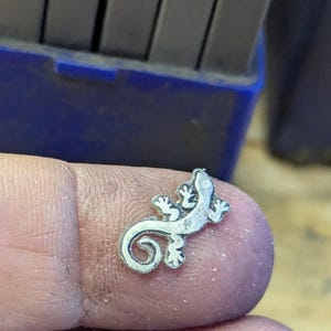 May include: A small, silver-colored lizard charm with a curled tail and detailed features. The charm is held on a finger, with a blue container and metal tools in the background. The lizard charm is a jewelry component.