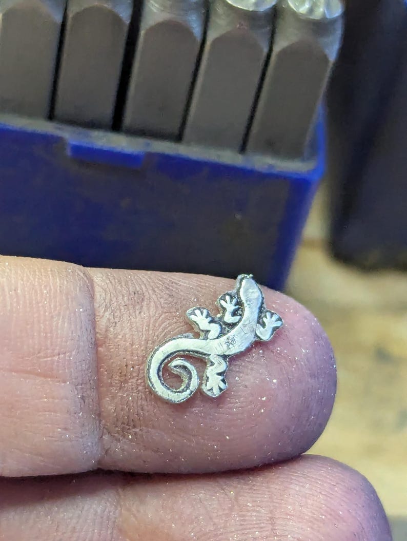 May include: A small, silver-colored lizard charm with a curled tail and detailed features. The charm is held on a finger, with a blue container and metal tools in the background. The lizard charm is a jewelry component.