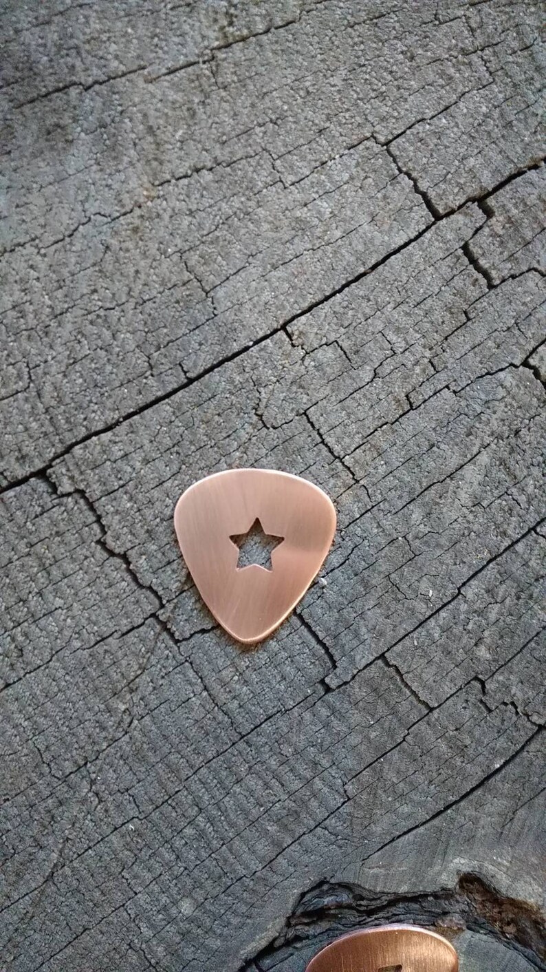 K&ouml;nnte beinhalten: Ein Kupfer-Plektrum mit einem Stern, der in der Mitte ausgeschnitten ist. Das Plektrum liegt auf einer Holzoberfl&auml;che.