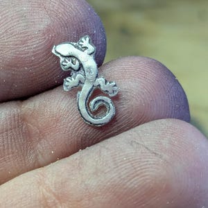May include: A small, detailed silver lizard charm with a curled tail. The lizard has four legs, each with three toes, and a textured body. The charm is likely a pendant or a component for jewelry making.