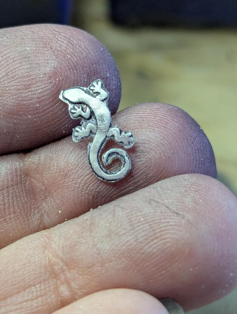 May include: A small, detailed silver lizard charm with a curled tail. The lizard has four legs, each with three toes, and a textured body. The charm is likely a pendant or a component for jewelry making.