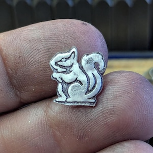 Op de afbeelding: Een kleine, zilverkleurige metalen eekhoornbedel. De bedel is gedetailleerd met ge&euml;tste lijnen, die de kenmerken van de eekhoorn laten zien, inclusief zijn volle staart en rechtopstaande houding. De bedel wordt in een hand gehouden, met een vage achtergrond.