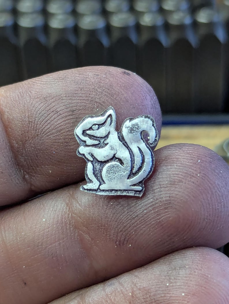Op de afbeelding: Een kleine, zilverkleurige metalen eekhoornbedel. De bedel is gedetailleerd met ge&euml;tste lijnen, die de kenmerken van de eekhoorn laten zien, inclusief zijn volle staart en rechtopstaande houding. De bedel wordt in een hand gehouden, met een vage achtergrond.