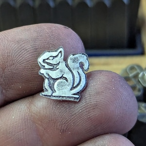 Op de afbeelding: Een kleine, zilverkleurige metalen eekhoornbedel. De bedel is gedetailleerd met ge&euml;tste lijnen, die de kenmerken van de eekhoorn laten zien. De eekhoorn zit met zijn staart opgerold. De bedel wordt in een hand gehouden.