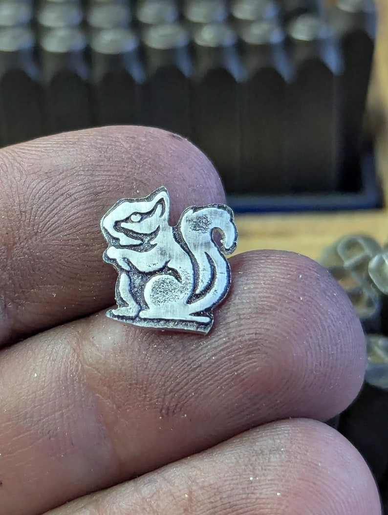 Op de afbeelding: Een kleine, zilverkleurige metalen eekhoornbedel. De bedel is gedetailleerd met ge&euml;tste lijnen, die de kenmerken van de eekhoorn laten zien. De eekhoorn zit met zijn staart opgerold. De bedel wordt in een hand gehouden.