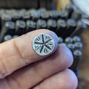 K&ouml;nnte beinhalten: Nahaufnahme eines Metallstempels mit floralem Design, der zwischen Fingern gehalten wird. Der Stempel hat eine kreisf&ouml;rmige Fl&auml;che mit einem f&uuml;nfbl&auml;ttrigen Blumenmuster. Weitere Metallstempel sind im Hintergrund sichtbar.