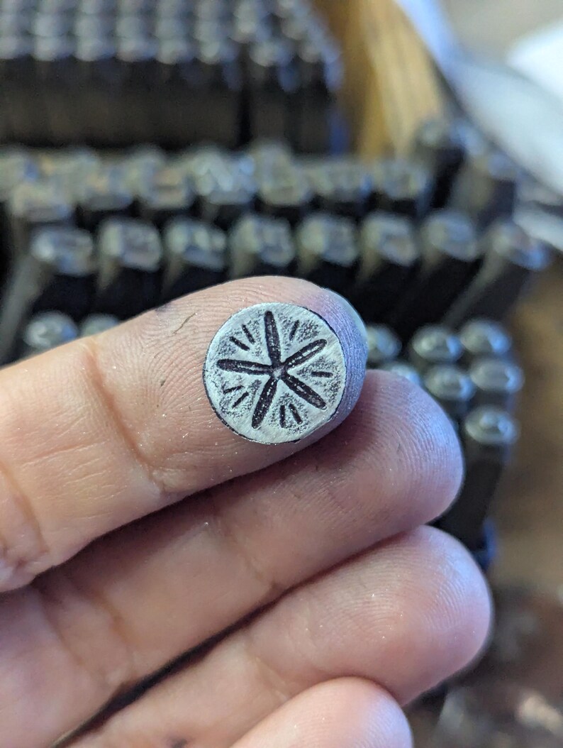 K&ouml;nnte beinhalten: Nahaufnahme eines Metallstempels mit floralem Design, der zwischen Fingern gehalten wird. Der Stempel hat eine kreisf&ouml;rmige Fl&auml;che mit einem f&uuml;nfbl&auml;ttrigen Blumenmuster. Weitere Metallstempel sind im Hintergrund sichtbar.