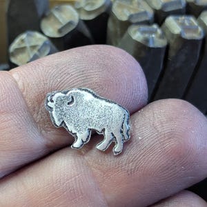 May include: A small, silver-colored metal charm in the shape of a bison. The charm is detailed with textured fur and stands on four legs. The background shows metal stamps.