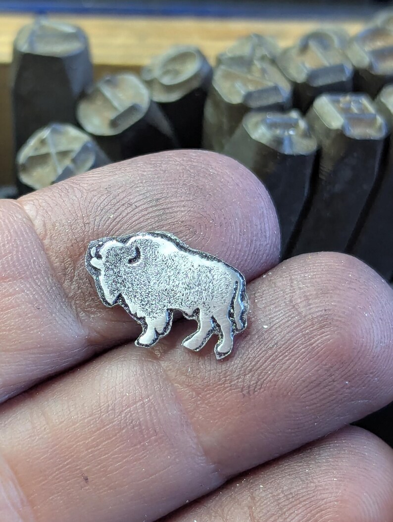 May include: A small, silver-colored metal charm in the shape of a bison. The charm is detailed with textured fur and stands on four legs. The background shows metal stamps.