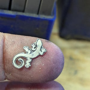 May include: A small, silver-colored lizard pendant with detailed features. The lizard has a curled tail and is resting on a finger. The background includes a blue container and metal tools.